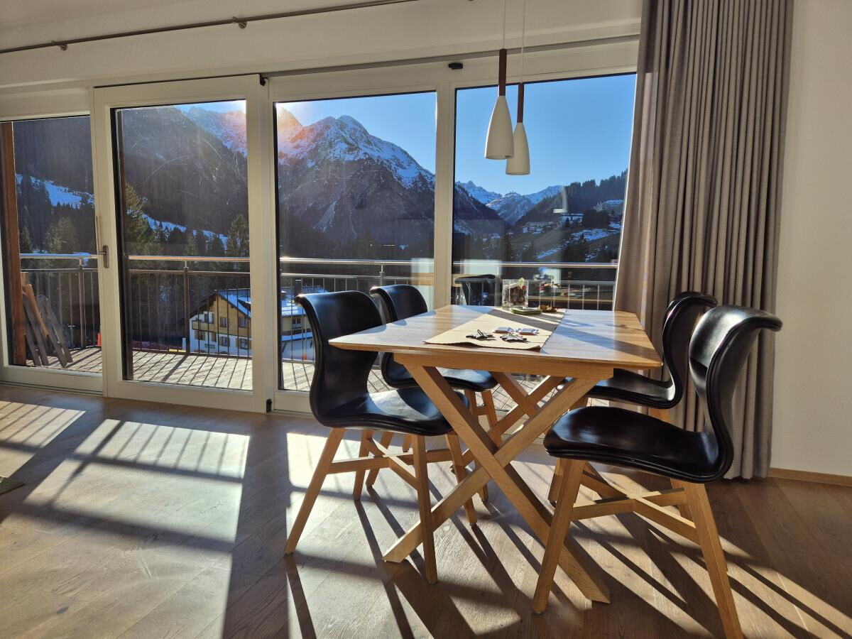 Dining table for six in front of the balcony windows with a view of the snow-covered mountains in the Sonnenbalkon luxury lodge Bärenkopf in the Kleinwalsertal
