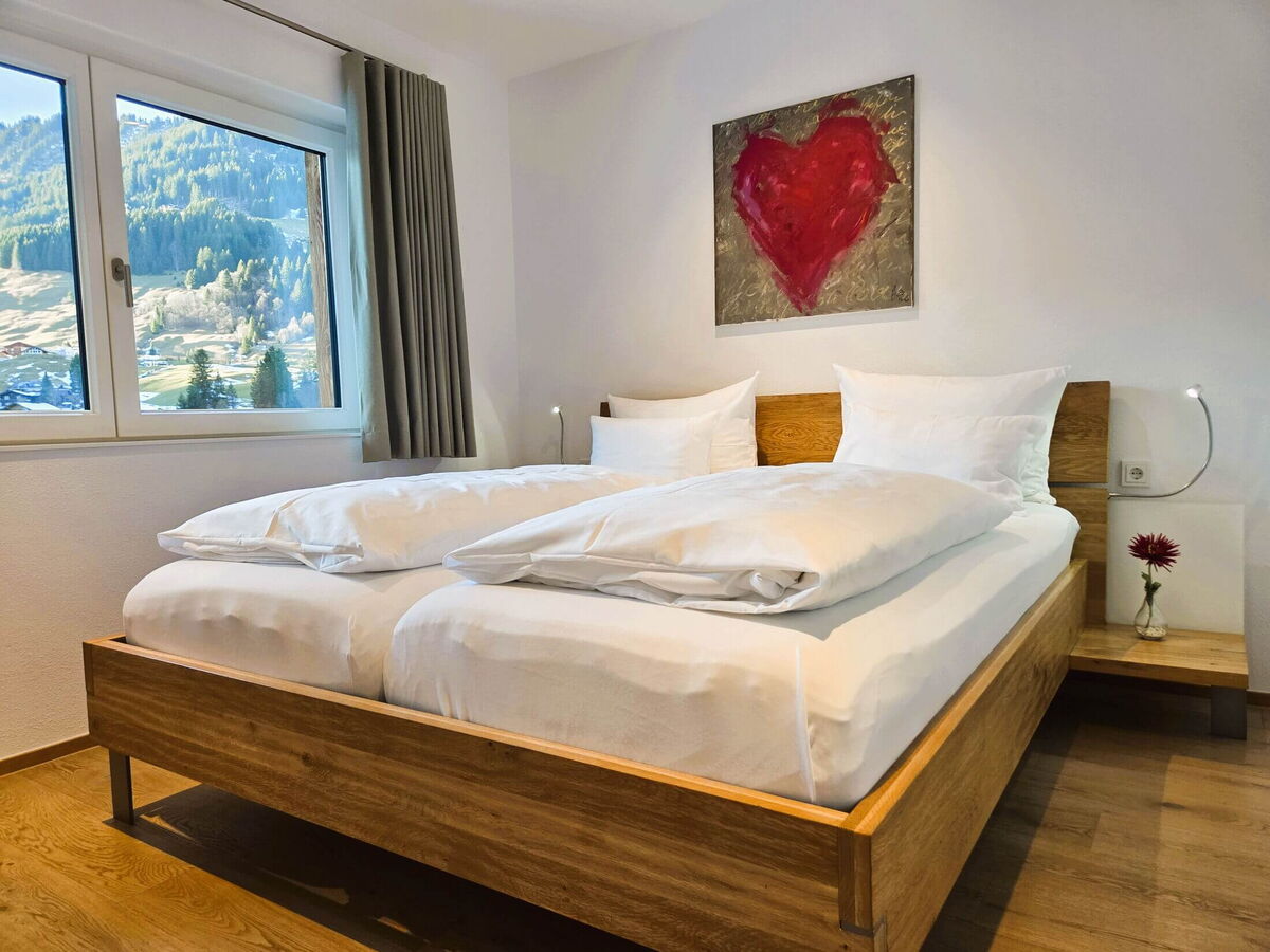 Schlafzimmer mit Massivholz-Doppelbett, rotem Herzbild und Fenster mit Blick auf die Alpenwiesen in der Bergwiese-Luxuslodge Zwölferkopf, Luxuslodge für 6 Personen im Kleinwalsertal