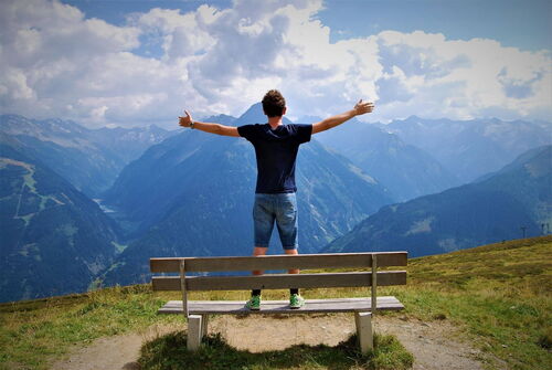Ein Mann steht auf einer Holzbank inmitten der Alpen, breitet die Arme aus und blickt auf die majestätischen Berggipfel vor blauem Himmel mit Wolken.