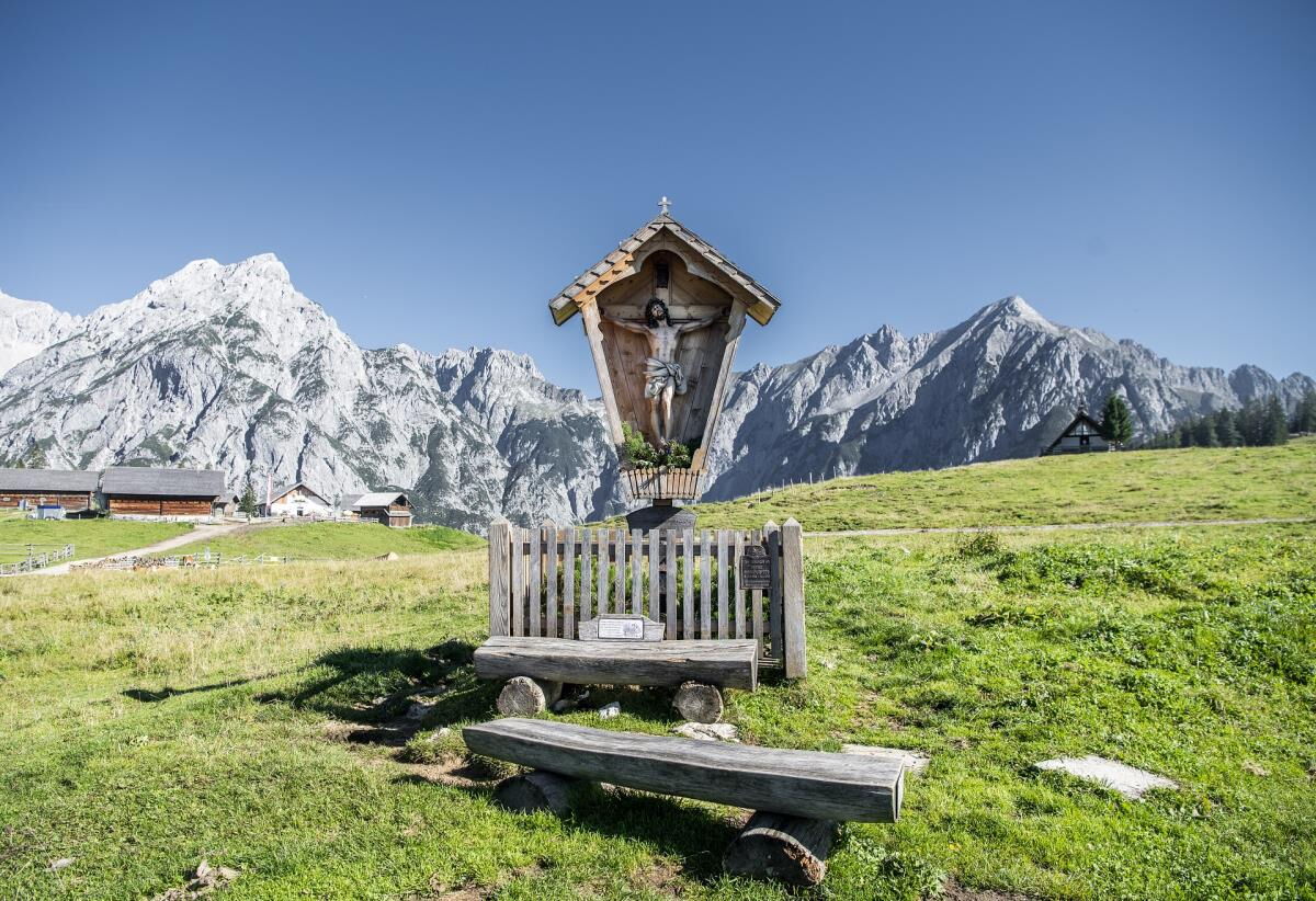 Hölzerner Bildstock mit Bank auf der grünen Wiese vor den verschneiten Bergen des Karwendels, Umgebung der Pferdeheimat am Karwendel in Gnadenwald