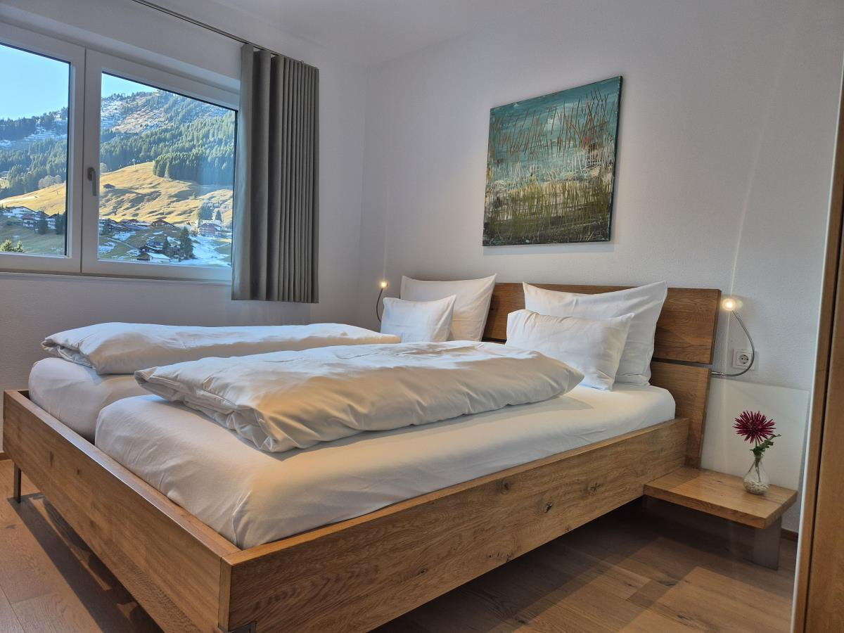 Bedroom with a solid wood double bed and a window onto the green slopes in the Sonnenbalkon luxury lodge Bärenkopf, holiday apartment for families in the Kleinwalsertal