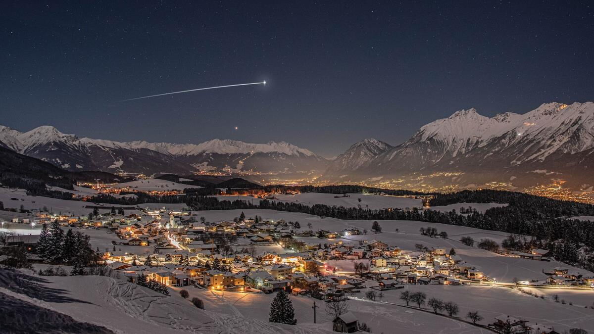 Winternacht über dem beleuchteten Inntal mit verschneiten Bergen und einer Sternschnuppe am Himmel