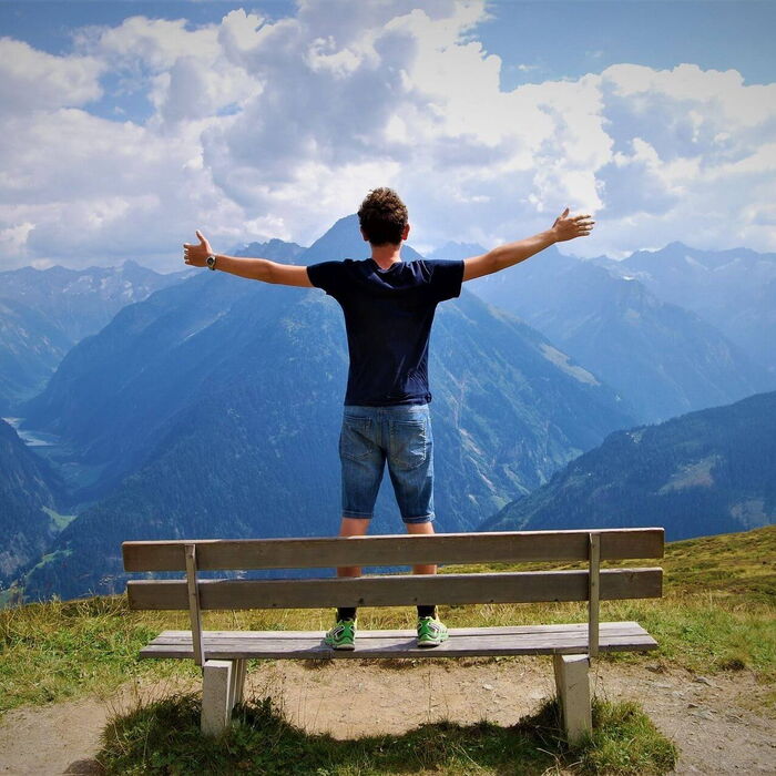Ein Mann steht auf einer Holzbank inmitten der Alpen, breitet die Arme aus und blickt auf die majestätischen Berggipfel vor blauem Himmel mit Wolken.
