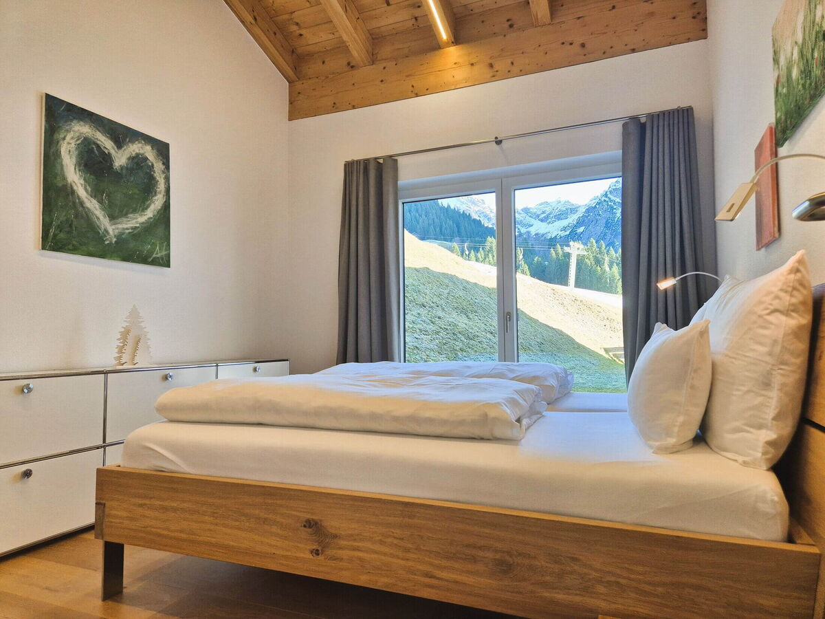 Bedroom with solid wood double bed, exposed roof beams and a floor-to-ceiling window onto the mountain meadow in the Panorama luxury lodge Ifen in the Kleinwalsertal