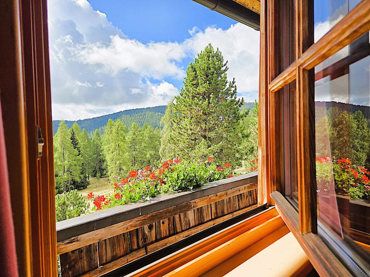 Blick aus dem offenen Holzfenster der Pietschacher Almhütte über rote Geranien auf Zirbenwald und die Nockberge