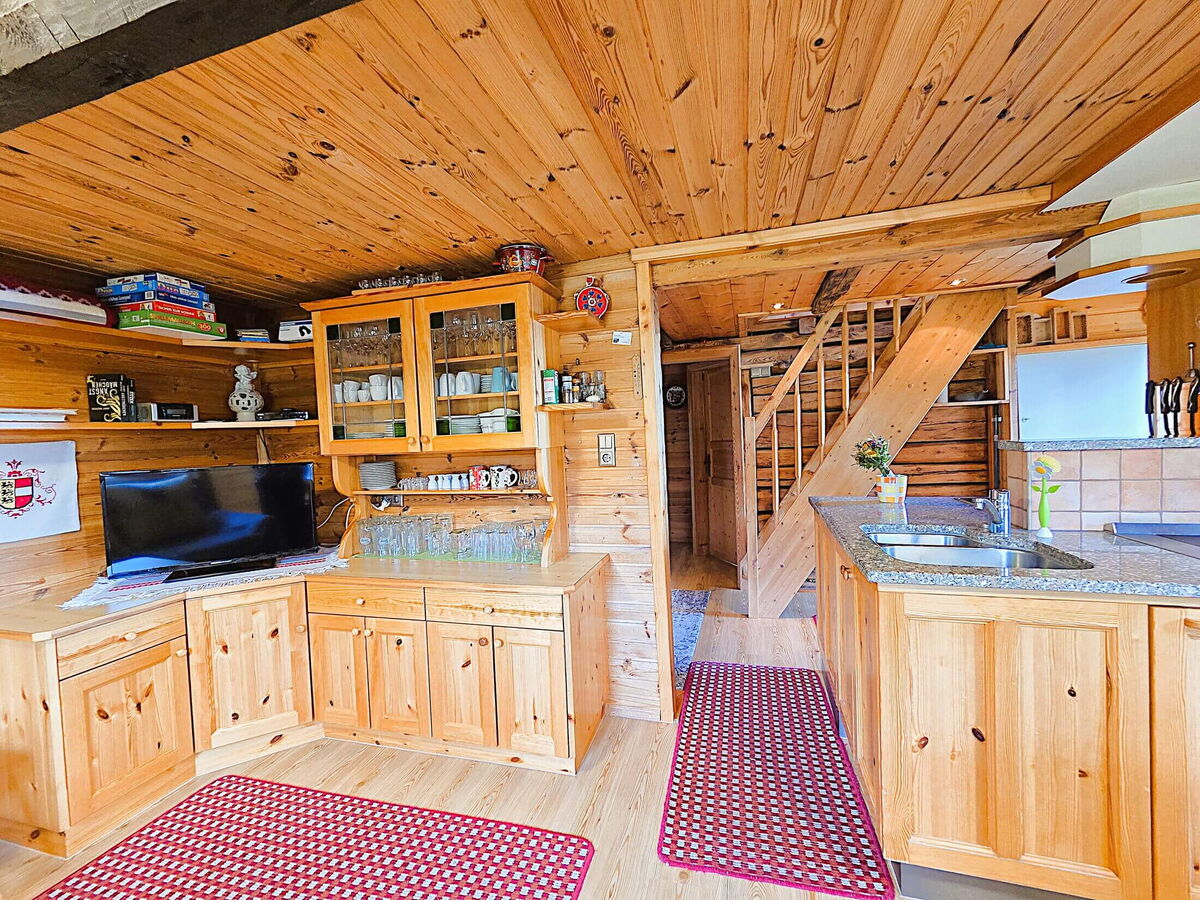 Rustikale Wohnküche der Pietschacher Almhütte mit Vitrine, Fernseher, Küchenzeile und Stiege ins Obergeschoss