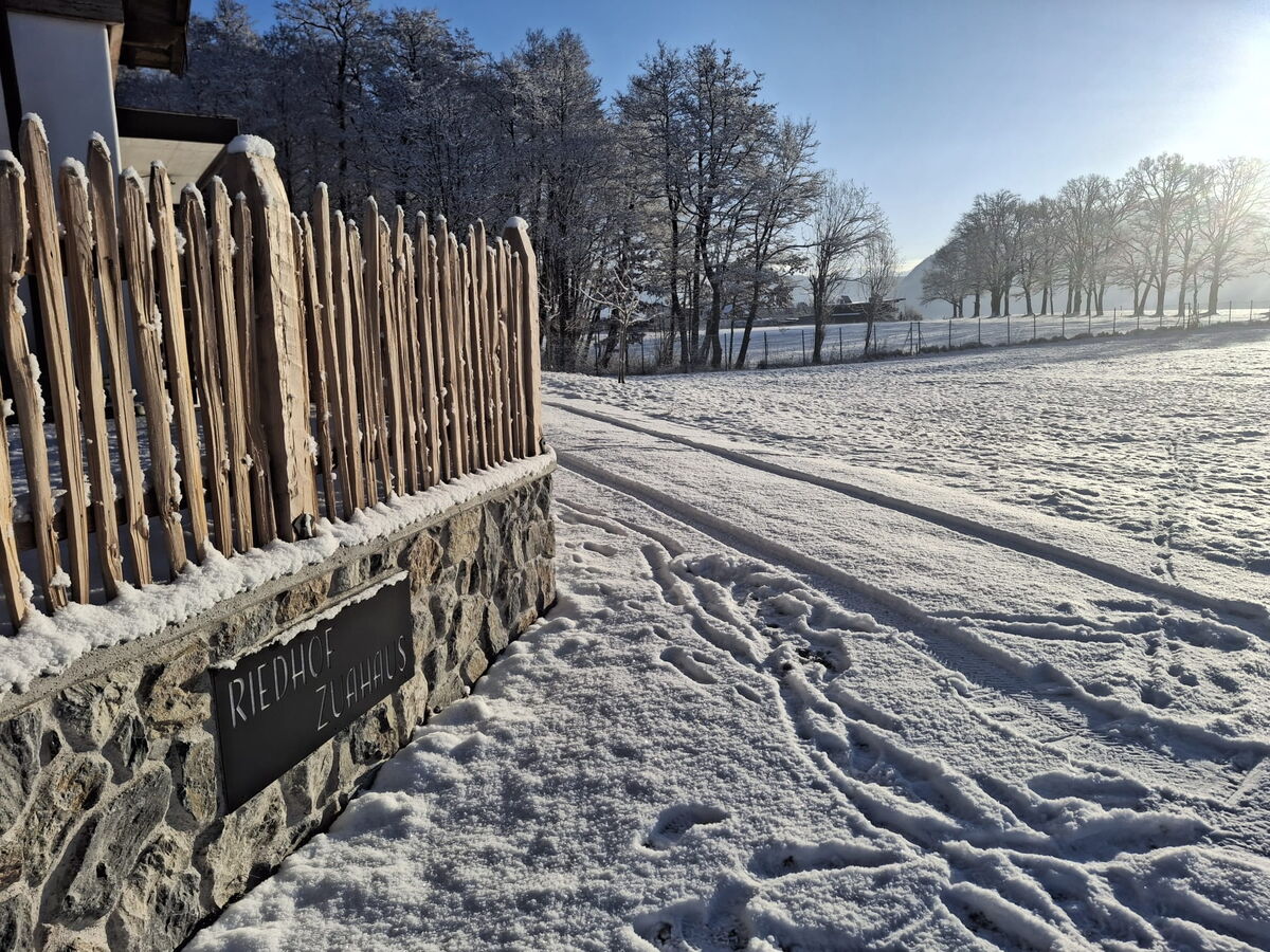 Holzzaun mit dem Schild Riedhof Zuahaus vor der verschneiten Wiese in Breitenbach, Winterstimmung am Bio-Bauernhof in Tirol