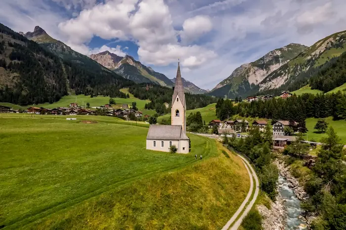 Matrei in Osttirol von oben: Kirche auf der Bergwiese, Ortsteile und Gebirgsbach vor den Hohen Tauern