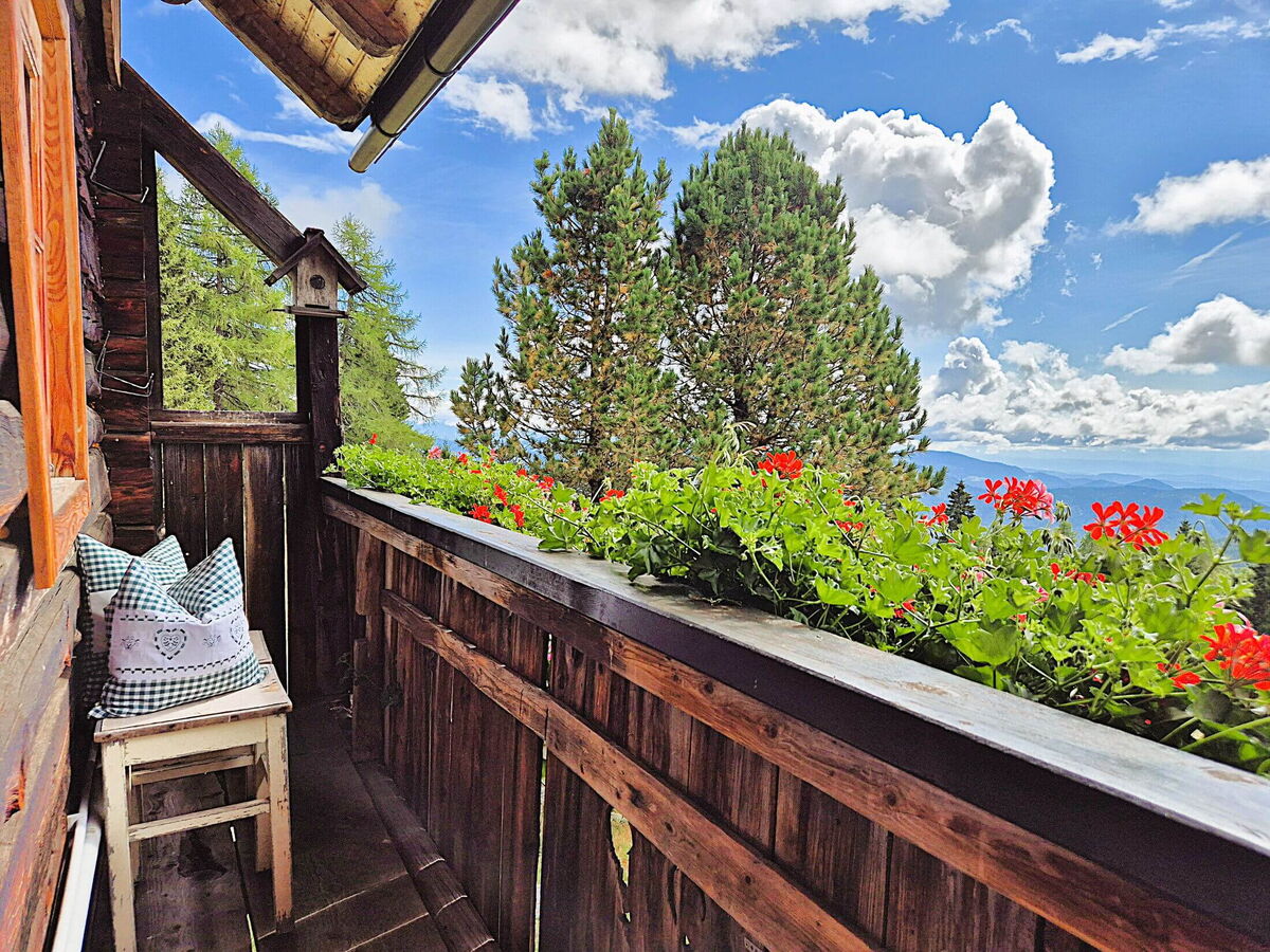 Balkon der Pietschacher Almhütte mit roten Geranien am Holzgeländer und Blick über Zirben und Wolken