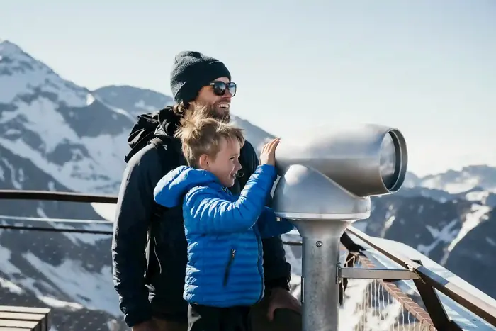 Father and child at the telescope on the viewing platform in front of the snow-covered glacier peaks