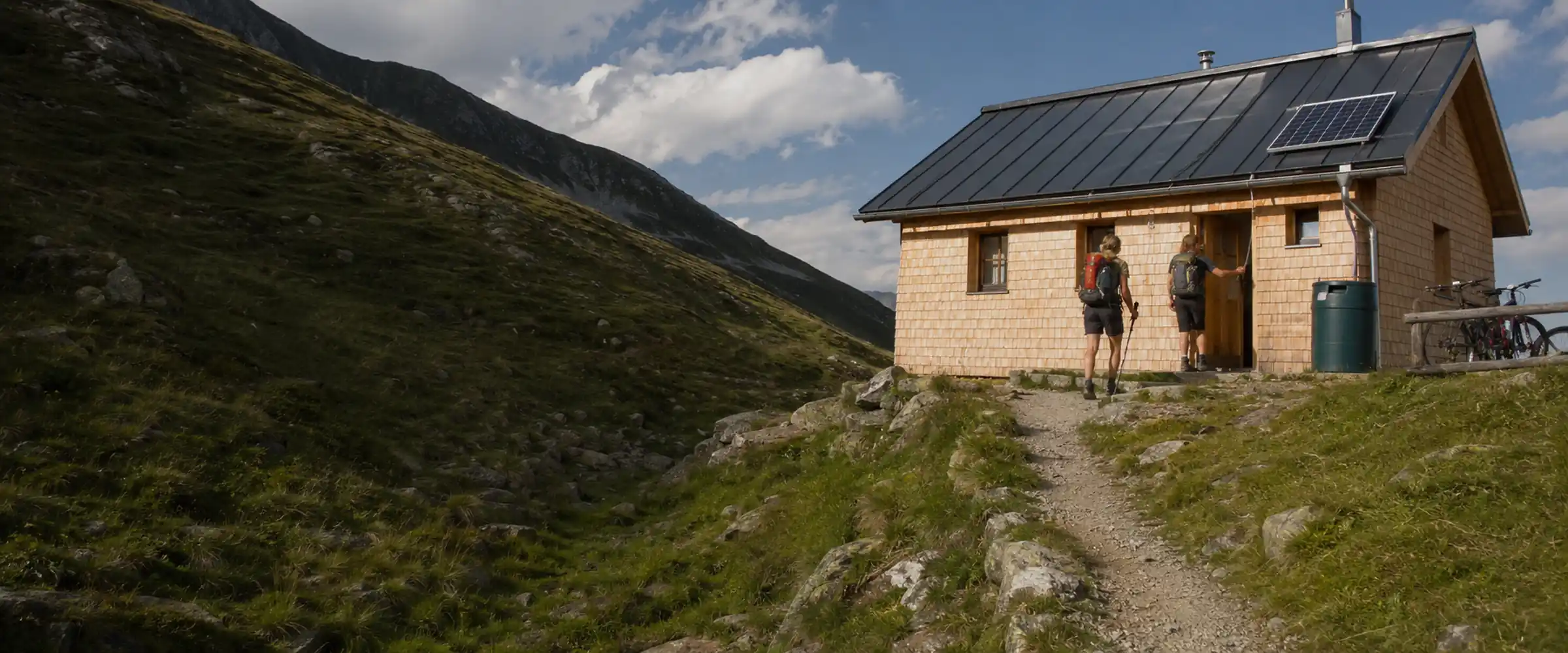 Two hikers reach an alpine hut with a solar panel and rainwater tank